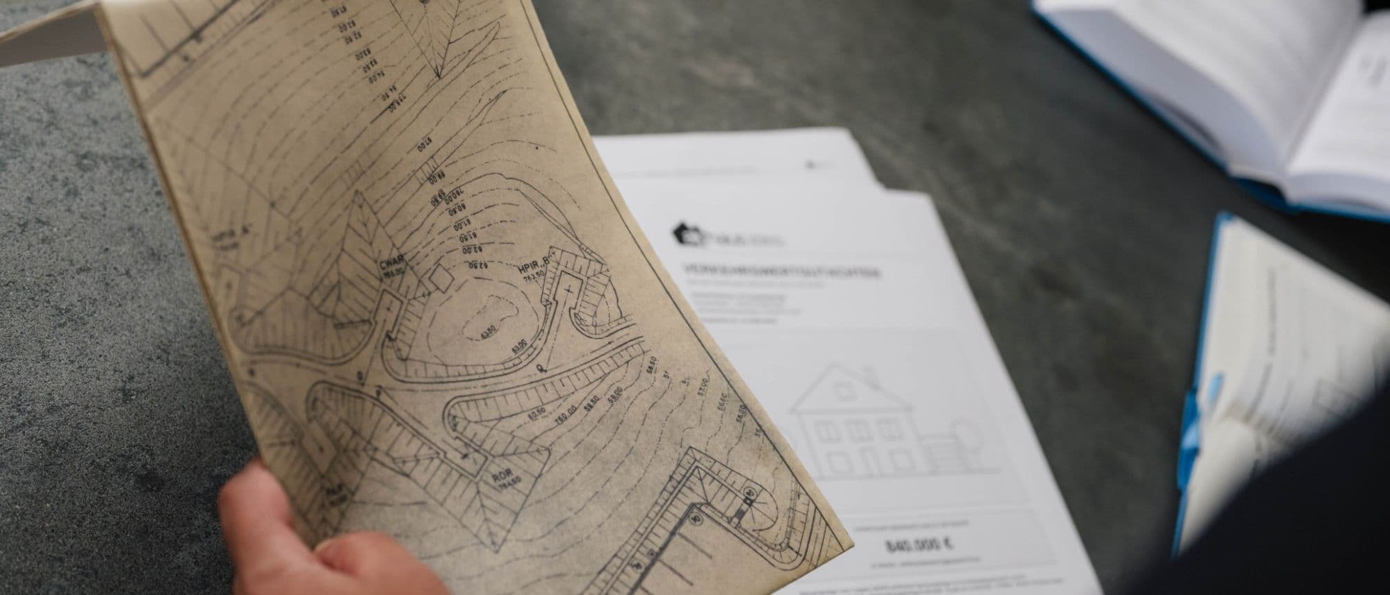 Lageplan des Grundstücks in der Hand, darunter das Deckblatt eines Verkehrswertgutachtens (Musterexemplar): Boden und Gebäude werden getrennt bewertet